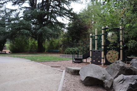 Playground on bark-chip surface &ndash; rocks to sit and climb on &ndash; bench and picnic table under tree on natural surface